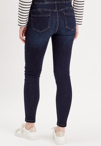 Jean skinny bleu foncé avec une texture denim lisse, taille haute et deux poches arrière, associés à des baskets blanches avec des accents métalliques.