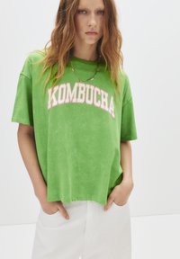 T-shirt verde oversize realizzata in materiale morbido, con la scritta "KOMBUCHA" in caratteri bianchi e rosa in grassetto sul petto. Abbinata a pantaloni bianchi.