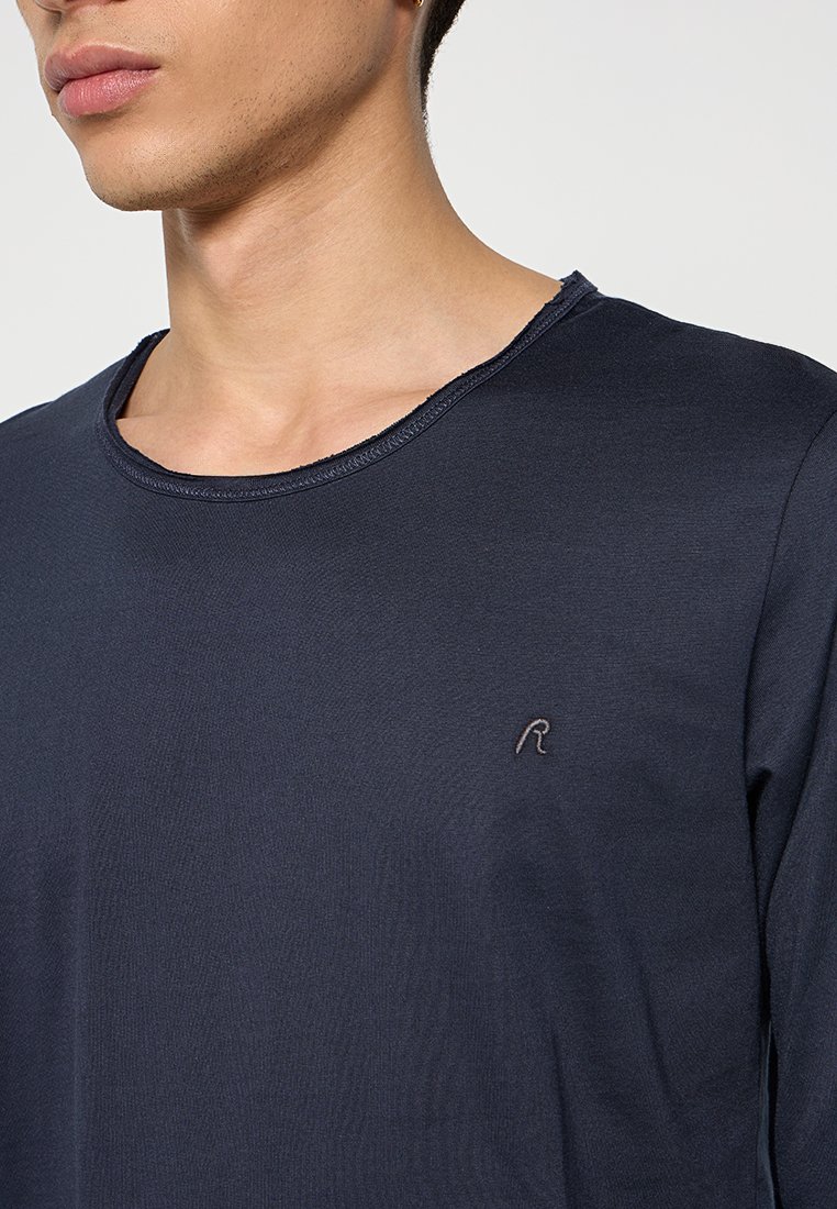 Junge Person in einem dunkelblauen Rundhalsausschnitt-Shirt mit einem kleinen gestickten "R" auf der Brust, gezeigt von den Schultern bis zu den Lippen.