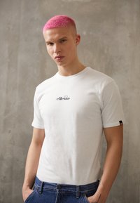 Weißes Baumwoll-T-Shirt mit einem schwarzen "ellesse"-Logo auf der Brust, kombiniert mit blauen Jeans, vor einem grauen Hintergrund präsentiert.