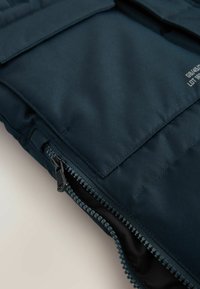Vesta din material navy cu închidere cu fermoar, având două buzunare cu clapetă și o suprafață texturată. Detaliu de cusătură de-a lungul fermoarului.