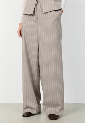 Beige broek met wijde pijpen en witte krijtstrepen, gedragen met donkerbruine schoenen met vierkante neus en een bijbehorende gilet met krijtstrepen.