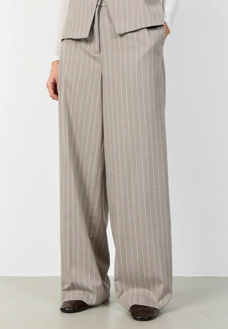 Pantaloni bej cu croială largă și dungi albe, purtați cu pantofi maro închis cu vârf pătrat și un veston asortat cu dungi albe.