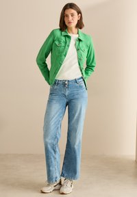 Veste en denim verte avec deux poches poitrine, haut blanc, jeans larges bleu clair, et baskets grises avec des accents. Fond uni.
