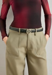 Cintura nera in pelle con una fibbia rotonda color oro caratterizzata da un design geometrico, indossata sopra pantaloni beige su misura con grandi tasche.