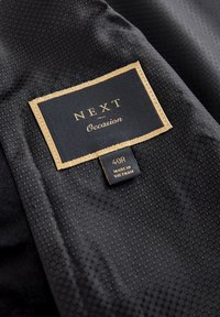 Tessuto nero con un motivo esagonale, caratterizzato da un'etichetta con accenti dorati che recita "NEXT Occasion" e la taglia "40R", prodotta in Vietnam.