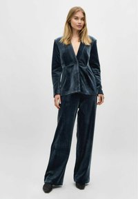 Marineblauwe fluwelen blazer met een geknipt revers en een enkele knoop, gecombineerd met bijpassende wijde broek. Zachte textuur overal.