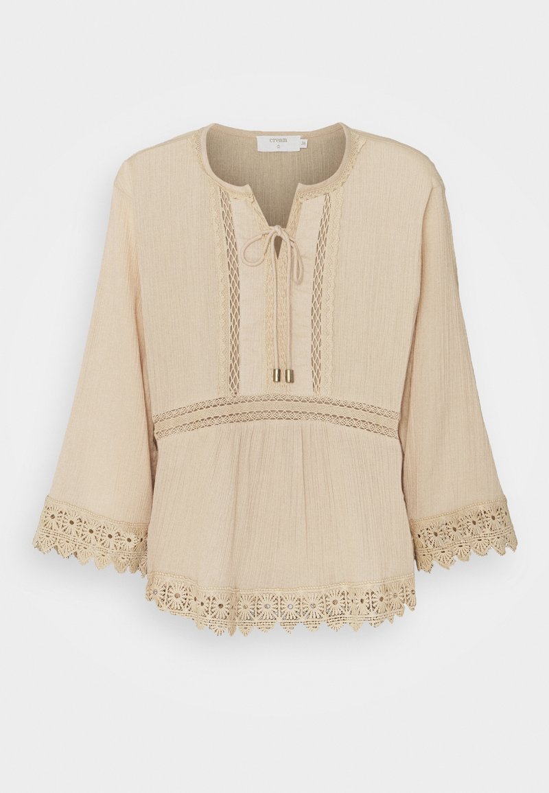 Blouse beige à manches longues avec bordure en dentelle aux poignets et à l'ourlet, panneaux de dentelle verticale et fermeture par lien au niveau de l'encolure ronde.
