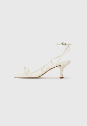 White heeled sandals with thin straps, square toe, and a 5 cm stiletto heel. Features an adjustable ankle strap with a metallic buckle.