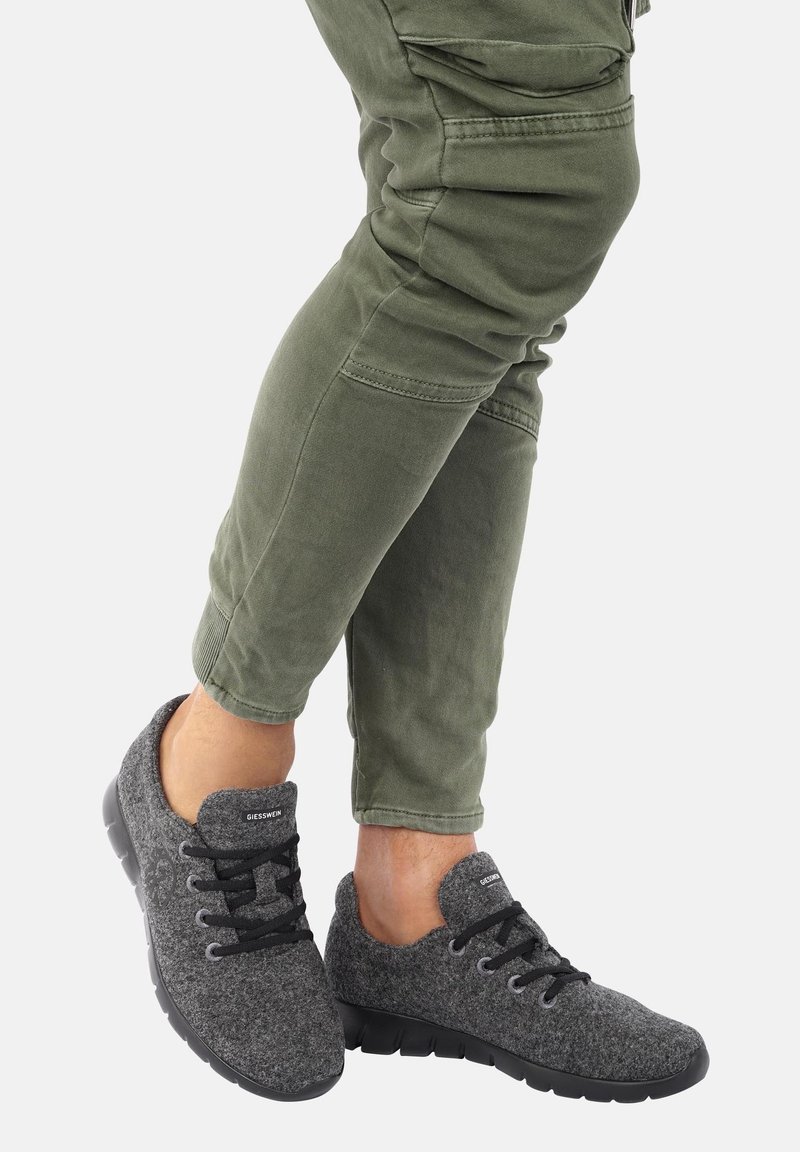 Personne portant un pantalon vert olive et des baskets basses grises avec des lacets noirs et des semelles noires flexibles, debout avec un pied légèrement levé.