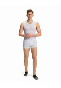 Débardeur blanc ajusté et boxer, mettant en valeur une texture lisse. Le modèle se tient pieds nus, avec des chaussettes foncées, devant un fond uni.