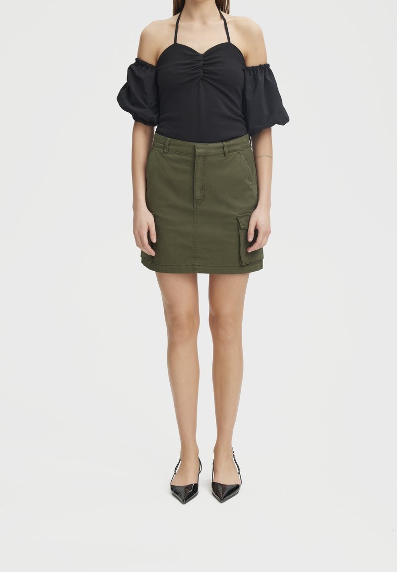 Femme portant un haut noir à manches bouffantes épaules dénudées avec col licou, jupe vert olive avec poche latérale et escarpins noirs à bouts pointus.