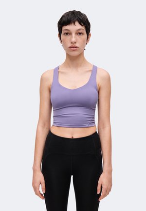 Mladá žena s krátkymi tmavými vlasmi, oblečená v priliehavom levanduľovom crop top a čiernych legínach s vysokým pásom, stojaca pred jednoduchým pozadím.