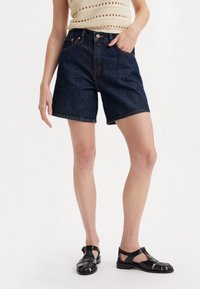 Denim shorts in dark blue, featuring a high waist and rolled hem. Paired with black, strappy sandals. Simple design, no visible patterns.