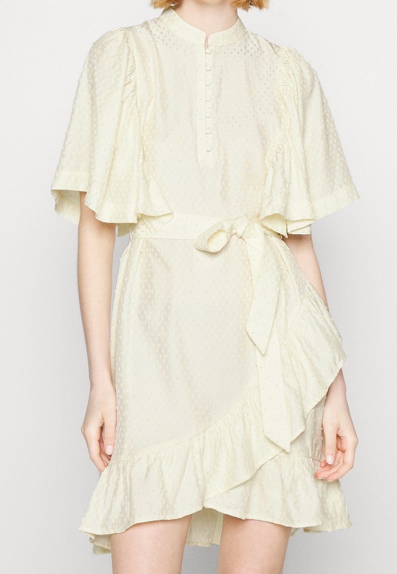 Robe courte à texture crème avec manches papillon, devant boutonné, ourlet à volants et ceinture nouée à la taille sur une femme debout.
