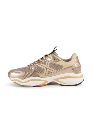 Zapatilla deportiva beige y dorado metálico con paneles de malla, lengüeta negra, cordones beige y suela gruesa blanca y beige.
