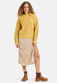Pull en laine jaune avec un col montant, jupe beige à motif floral avec une fente cuisse, et bottines brunes à empiècements élastiques.