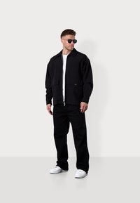 Black zip-up jacket with oversized pockets, paired with black pants and white sneakers. The model wears sunglasses and a white T-shirt underneath.