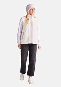 Fleece vest in off-white with lavender accents, zip closure, two side pockets, paired with a lavender sweater, black jeans, and white sneakers.