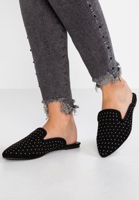 Svarta mockaskor utan snörning med spetsiga tår, dekorerade med silverstift, i kombination med grå fransade jeans med dekorativa knappar.