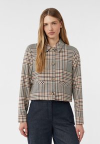 Žena v krátké, knoflíkované kostkované bundě s dlouhými rukávy a tmavých kalhotách s vysokým pasem, stojící na jednobarevném pozadí.