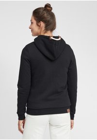 Black hoodie with a drawstring at the neck, ribbed cuffs, and a leather accent at the hem, worn over light-colored pants.