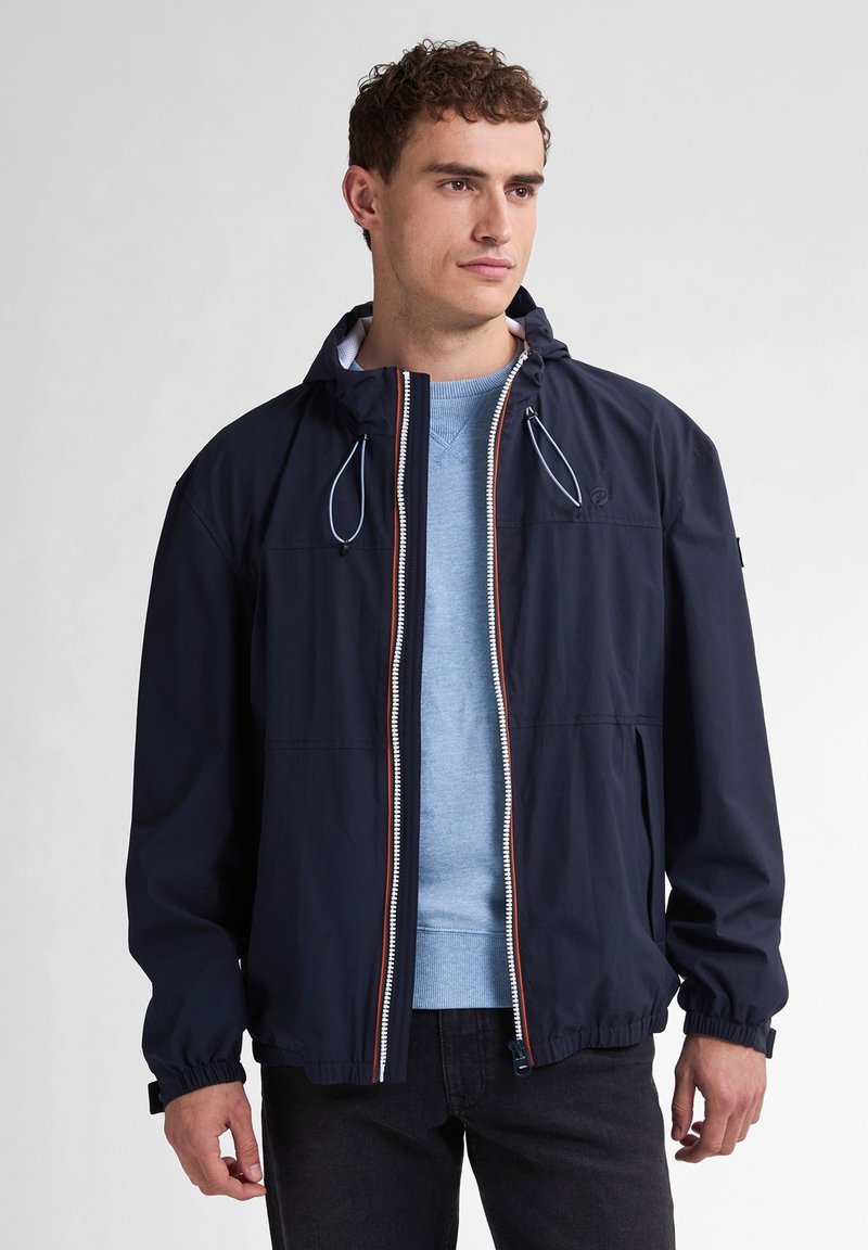 Marine windbreakerjack met elastische manchetten, volledige rits en verstelbare capuchon. Gedragen over een lichtblauw sweater en donkere jeans.