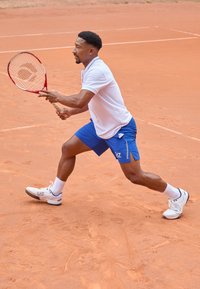 Teniszjátékos fehér pólóban és kék sortban, piros teniszütőt használva a salakpályán, fehér sportcipőkben, amelyek logóval rendelkeznek.