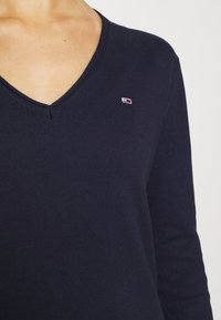 Gros plan d'une personne portant un pull à col en V bleu marine avec un petit logo rectangulaire rouge, blanc et bleu sur la poitrine.