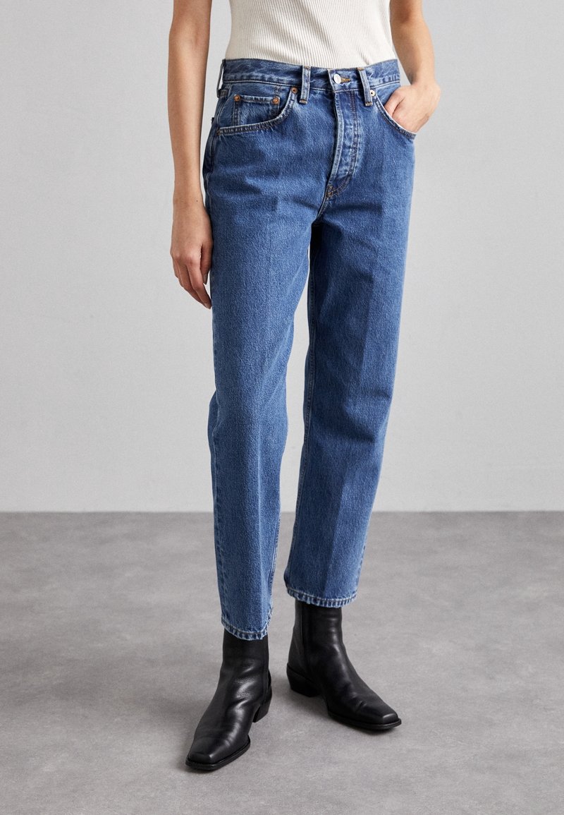 Personne portant un jean bleu droit taille haute, un haut blanc sans manches et des bottines noires, debout sur un sol gris.