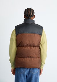 Gevoerde vest met een bruine body en een zwarte nylon bovenkant, hoge kraag en mouwloos ontwerp, gedragen over een lichtgroen langslavenshirt.