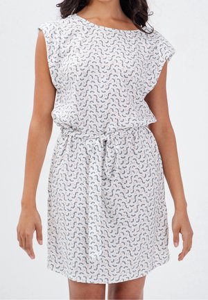 Nainen pukeutuneena valkoiseen lyhythihaiseen mekkoon, jossa on mustia ja punaisia abstrakteja kuvioita, vyötettynä vyötäröltä ohuella kangasvyöllä.