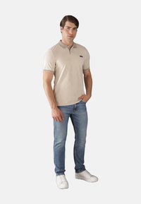 Polo beige con colletto e polsini a costine, abbinato a jeans azzurri chiari e sneakers bianche. Design semplice e vestibilità standard.