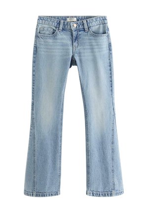 Ung person iført en hvid top med korte ærmer, lyseblå vide jeans og hvide sneakers mod en ensfarvet baggrund.