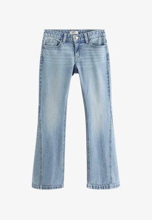 Ung person iført en hvid top med korte ærmer, lyseblå vide jeans og hvide sneakers mod en ensfarvet baggrund.