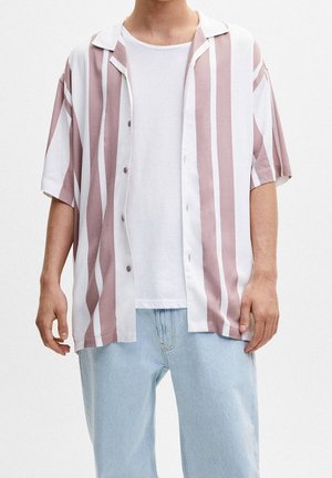 Homme portant un jean bleu clair, un t-shirt blanc et une chemise à manches courtes ouverte avec des rayures verticales roses et blanches.