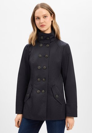 Woman wearing a black double-breasted coat with six buttons and two flap pockets, standing against a plain white background.