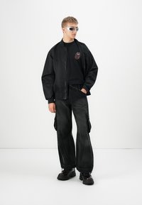 Młody mężczyzna w czarnej bomberce, czarnym t-shircie z grafiką, luźnych czarnych spodniach cargo, masywnych czarnych butach i srebrnych sportowych okularach przeciwsłonecznych.