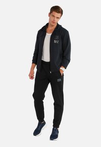 Felpa navy con zip e logo, t-shirt bianca, pantaloni jogger neri con patch logo, e scarpe da ginnastica navy con suole bianche. Abbigliamento sportivo casual.