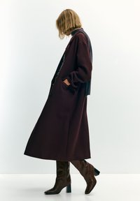 Vrouw met kort blond haar die een lange donkerbordeauxrode jas, bruine laarzen met hak en een marineblauwe sjaal draagt, lopend tegen een effen witte achtergrond.