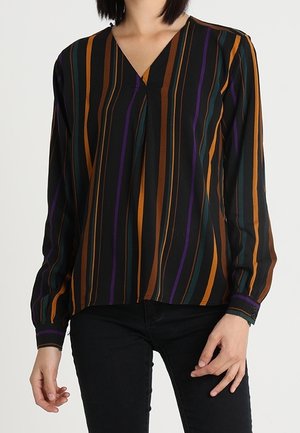 Femme portant un chemisier noir à manches longues avec des rayures verticales orange, marron, verte et violette et un pantalon noir.