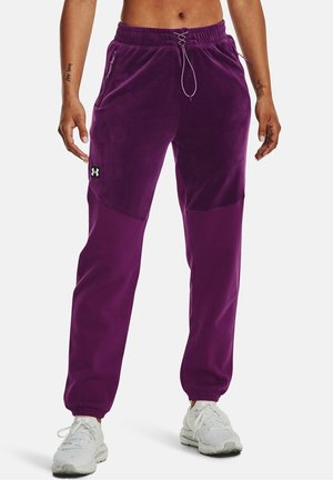 Personne portant un jogging sportif violet avec cordon de serrage et poches, associé à des chaussures de course blanches, debout devant un fond blanc.
