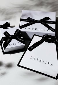 Trzy białe pudełka prezentowe z czarną wstążką, z logo "LATELITA". Różne rozmiary, gładka powierzchnia, podkreślone kontrastującymi czarnymi krawędziami.