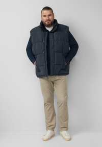 Granatowa pikowana kamizelka z wysokim kołnierzem, zapinana na guziki, z bocznymi kieszeniami, noszona na granatowej koszuli z długim rękawem i beżowych spodniach.