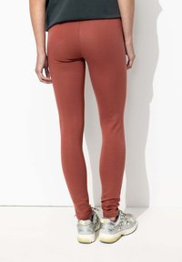 Leggings rouges extensibles en matériau lisse, présentant un design épuré qui se resserre à la cheville, associés à des baskets grises.
