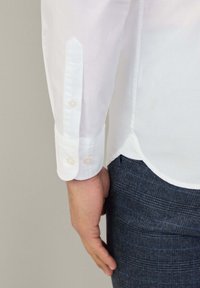 Camicia bianca in cotone con polsini a punta e due bottoni perla, abbinata a pantaloni grigio scuro a trama testurizzata con un sottile motivo a quadri.