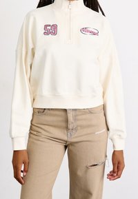 Felpa zip con cappuccio color crema, dotata di ricamo "59" e logo "ellesse", con una vestibilità rilassata e polsini a coste. Indossata con jeans beige strappati.