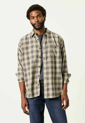 Man with beard wearing beige and gray checkered shirt over black t-shirt and blue jeans standing against plain light background.