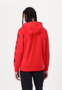 Röd bomullshoodie med en känguruficka och huva. Har svarta grafiska mönster på båda ärmarna samt ribbad nederkant och ärmslut.