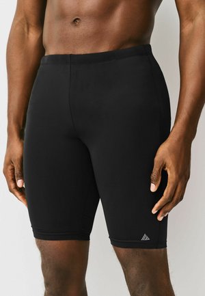 Muskuløs person iført tætsiddende sorte træningsshorts, stående mod en ensfarvet baggrund med hænderne afslappet langs siden.